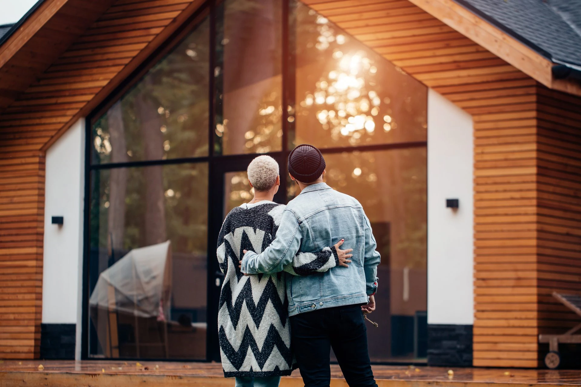 A young couple looks toward a cabin. A reflection of the sunset can be seen on the cabin's front windows.
