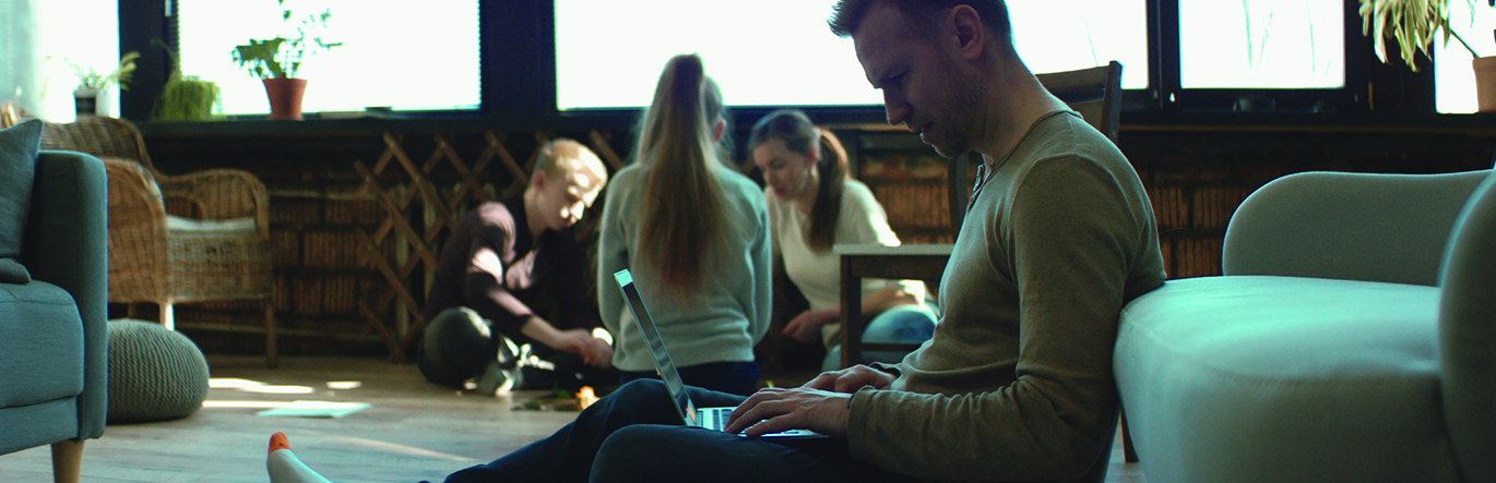 A father looks down at computer screen with concern while his family enjoy a board game in the background.