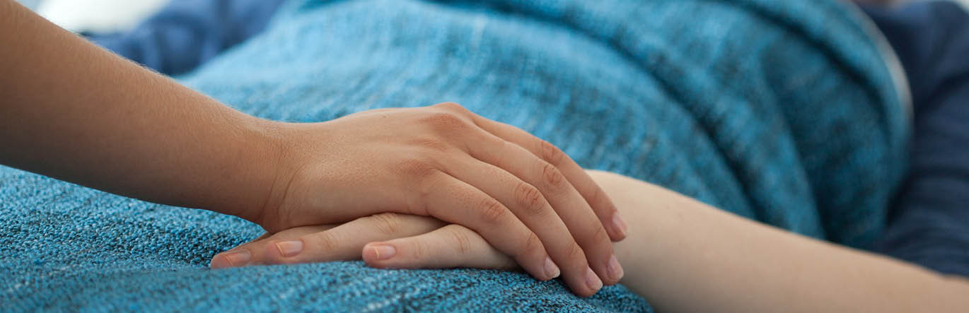 The hand of an ill person laying in bed is held by another.
