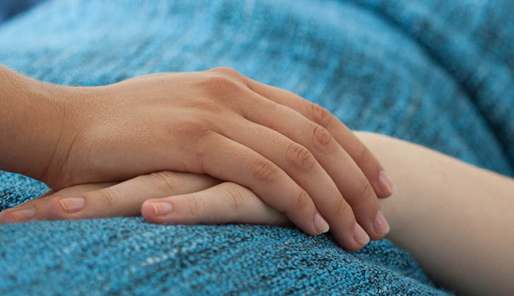 The hand of an ill person laying in bed is held by another.