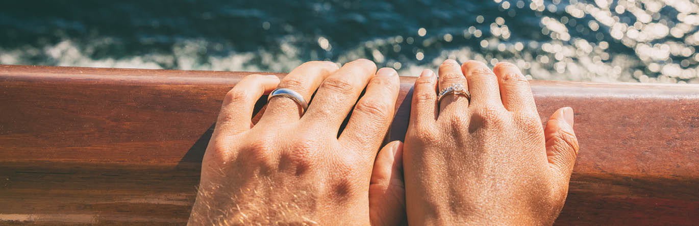 The hands of two individuals on a railing with wedding rings. There is moving water in the background.
