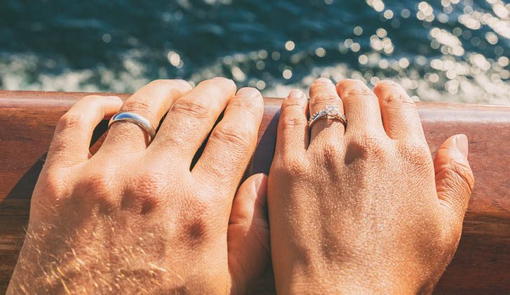 The hands of two individuals on a railing with wedding rings. There is moving water in the background.