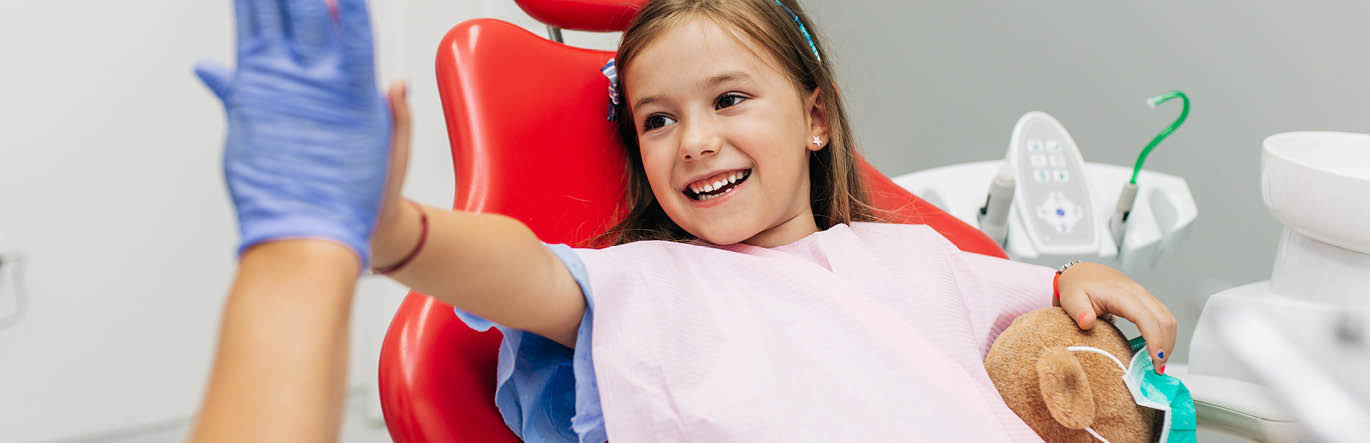 Child in a dentist chair, high-fiving a gloved hand with a smile in her face.