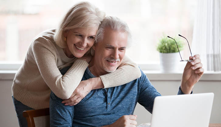 Older couple, husband sitting down while wife fondly hugs him from behind, review a computer screen.