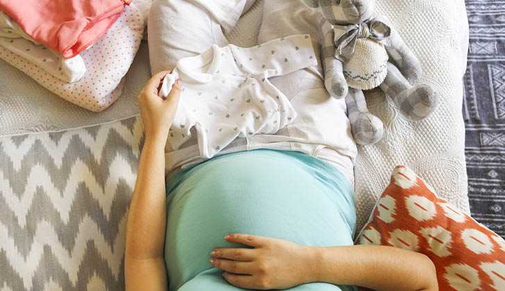 A pregnant mother caresses her bump, baby clothes around her in the bed.