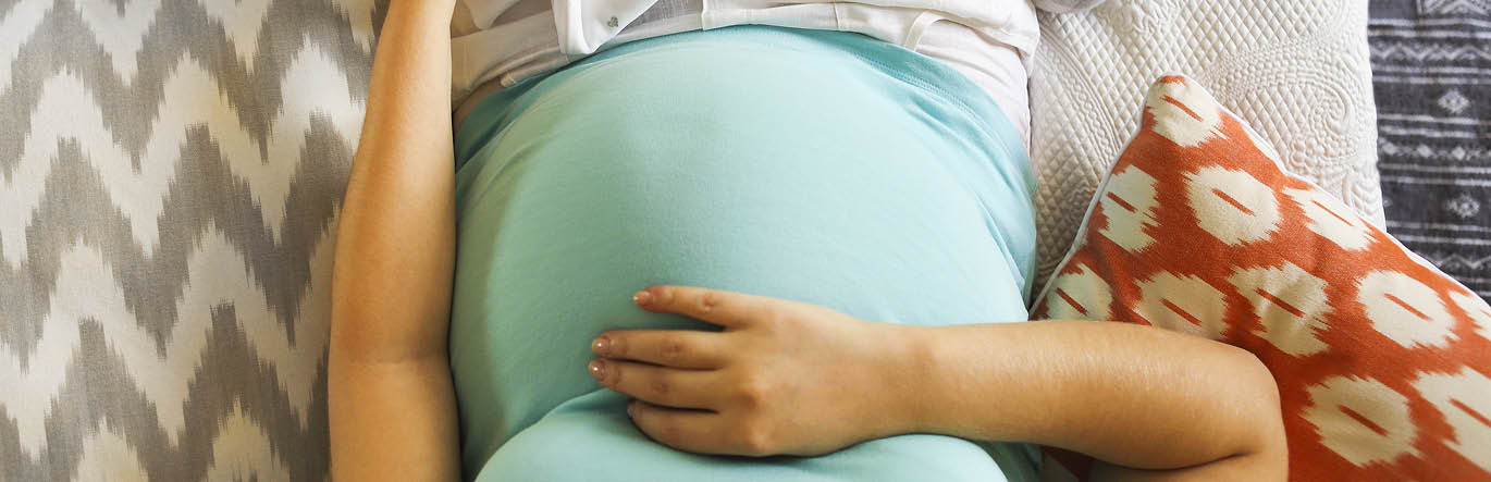 A pregnant mother caresses her bump, baby clothes around her in the bed.