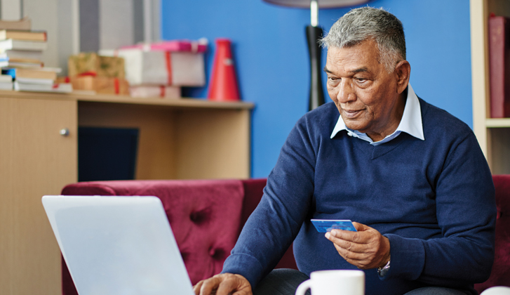 Gentleman shopping online, holding his financial card in one hand inputs the details on the computer with his other hand.