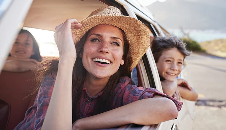 A mother looks out a window, while her children in the back seat smile at the camera.