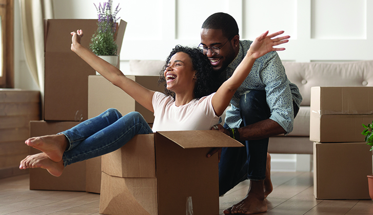 A young couple smiles as the husband pushes the wife who is inside a cardboard box.