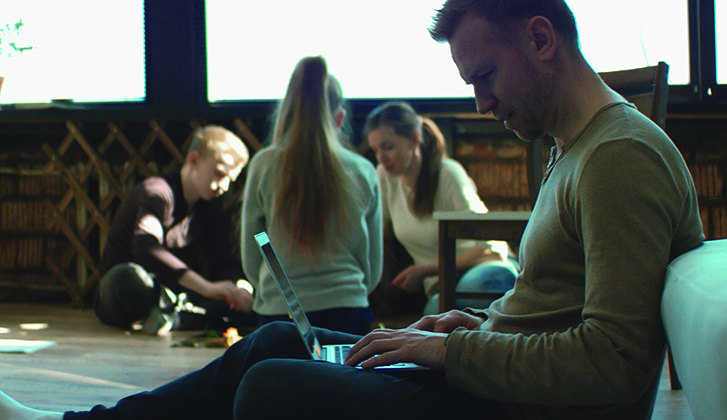 A father looks down at computer screen with concern while his family enjoy a board game in the background.