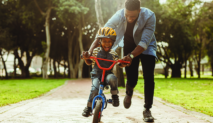 A smiling child learns to ride a bicycle while his parent pushes him along.