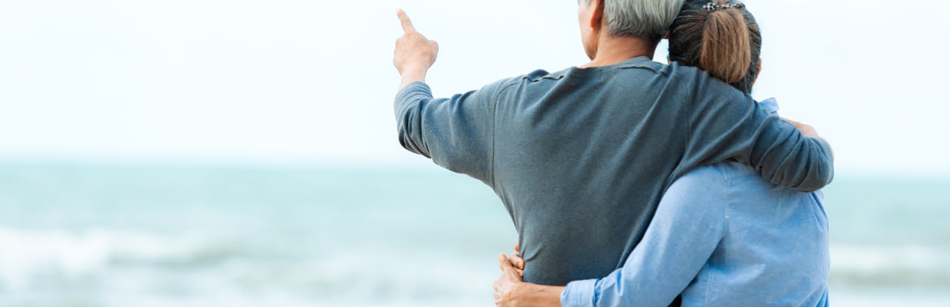 A man with grey hair hugs his wife as they look on a shore.