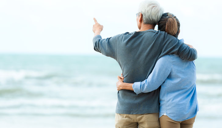 A man with grey hair hugs his wife as they look on a shore.