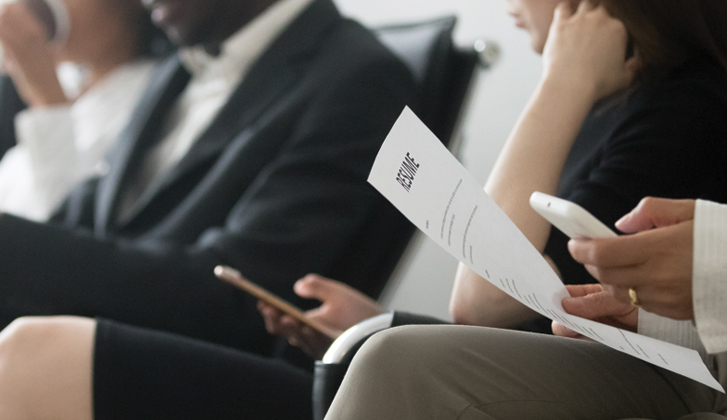 Perspective shot of group of well-dressed professionals sitting in a row in a waiting room with resumes in hand.