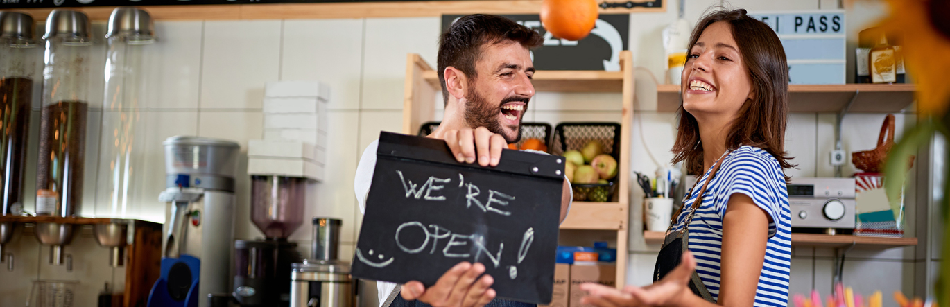 Business-owning couple with excited smiles holding a sign that says We're Open