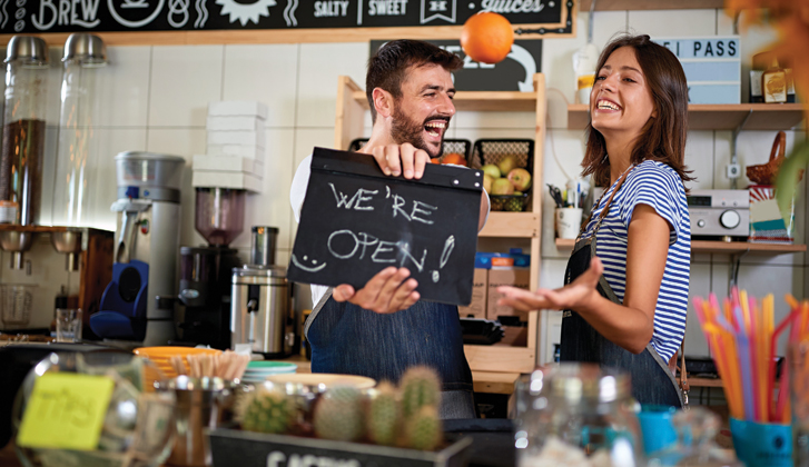 Business-owning couple with excited smiles holding a sign that says We're Open