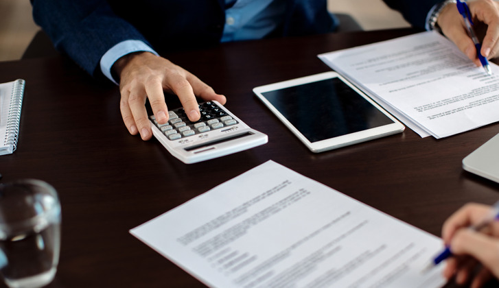 papers, calculators and a tablet on a desk. Two individuals work over the documents.