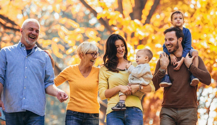 Grandparents, parents, and two children in their arms walk through autumn foliage.