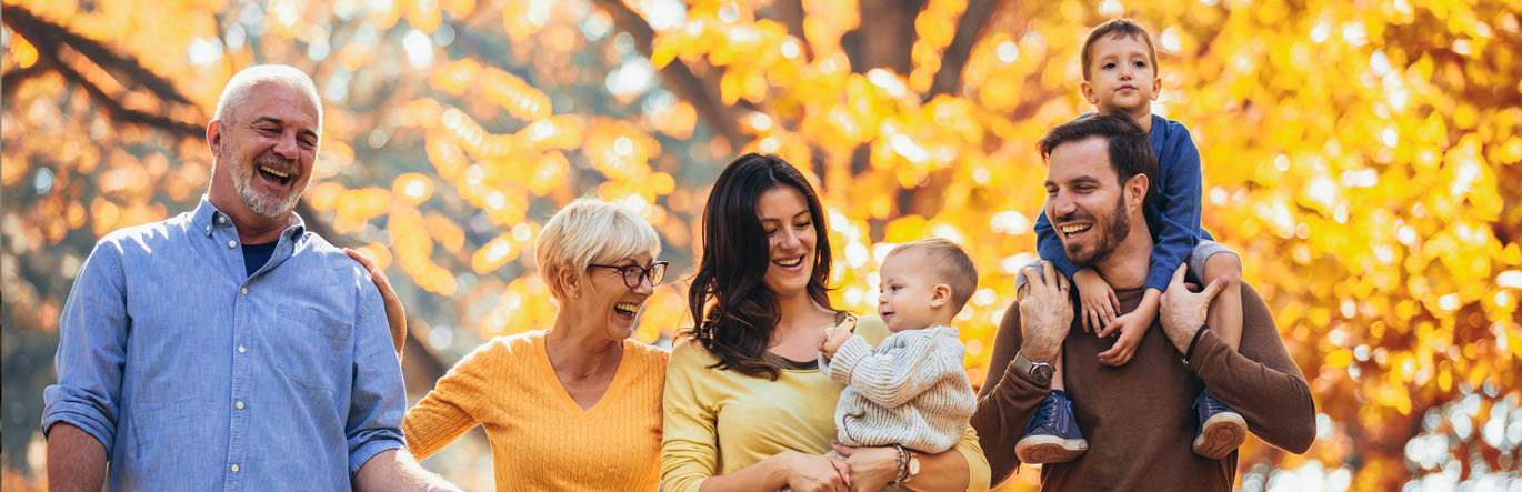 Grandparents, parents, and two children in their arms walk through autumn foliage.
