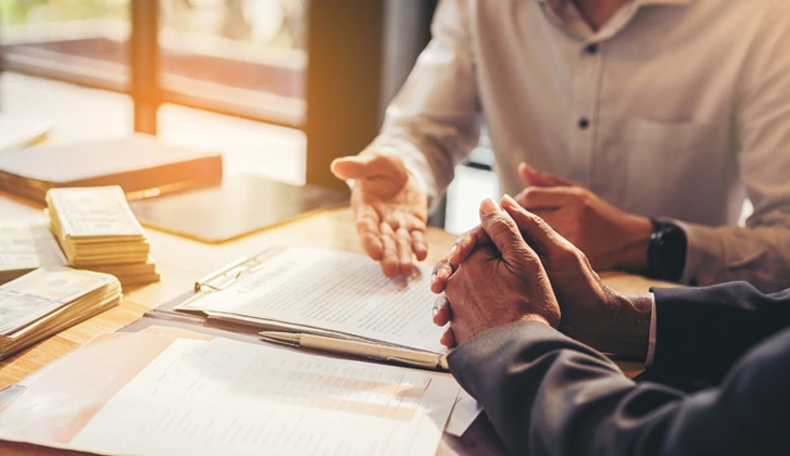 Gentleman with folded hands over a portfolio of documents receives guidance from other gentleman at his side.