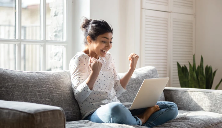 Young woman sitting on couch with computer on her lap smiles excitedly, pumping her fists in the air in celebration.