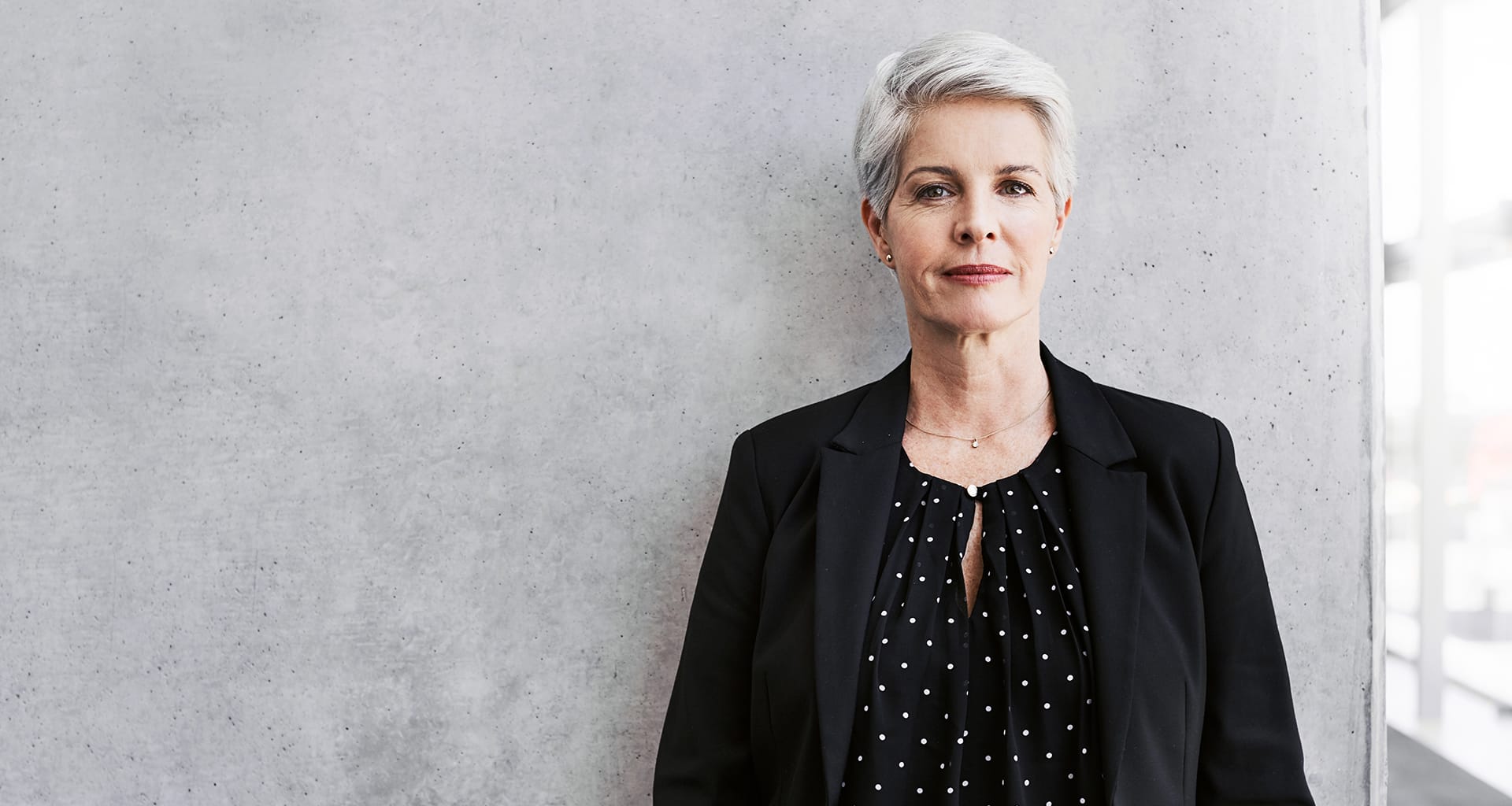 Slender woman with grey hair standing next to a window.