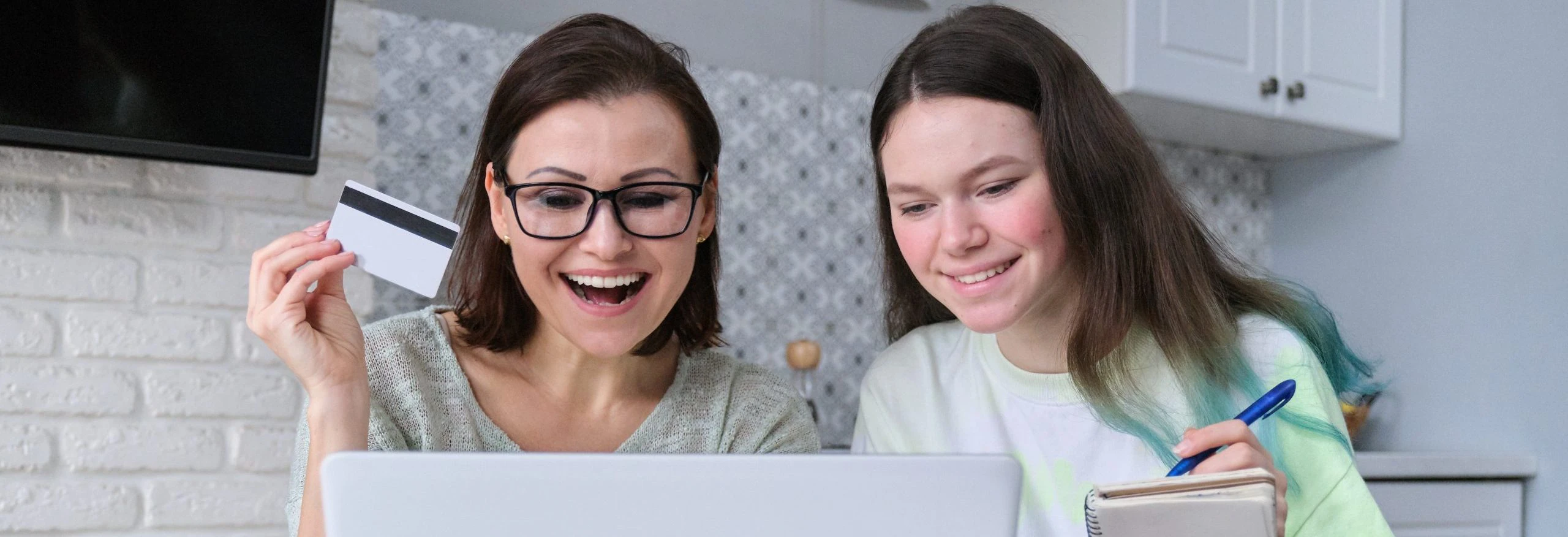 Mother is holding a debit card and her daughter is taking notes while they look at a laptop.