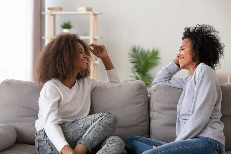 African American mother and daughter sit on sofa laughing and communicating facing each other.