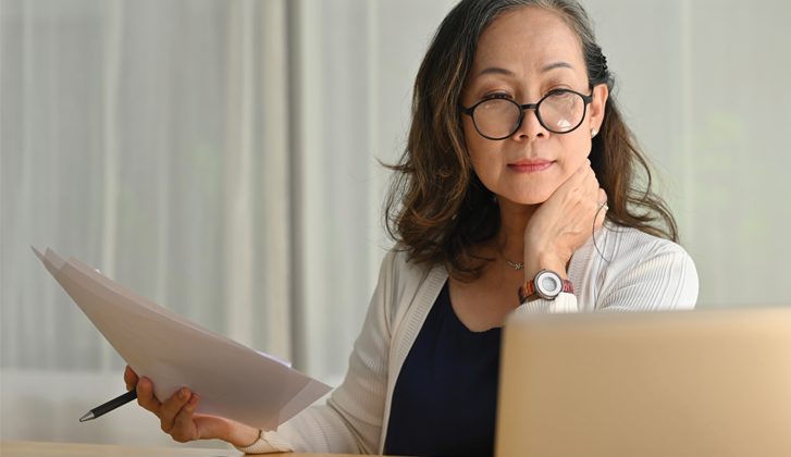 Older Asian woman holding documents while looking at a laptop screen.