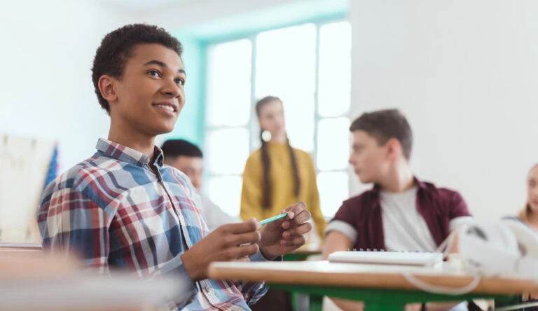 black highschool boy sitting at desk holding pencil with classmates in the background