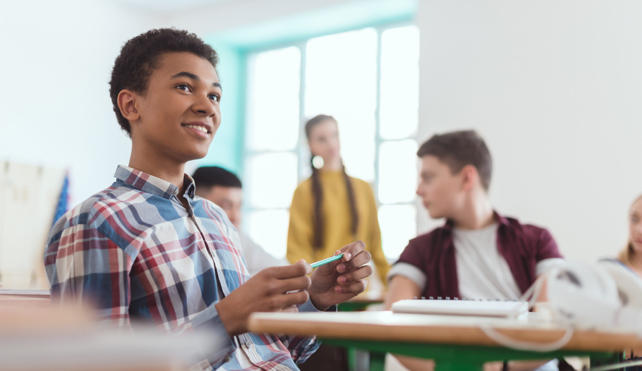 black highschool boy sitting at desk holding pencil with classmates in the background