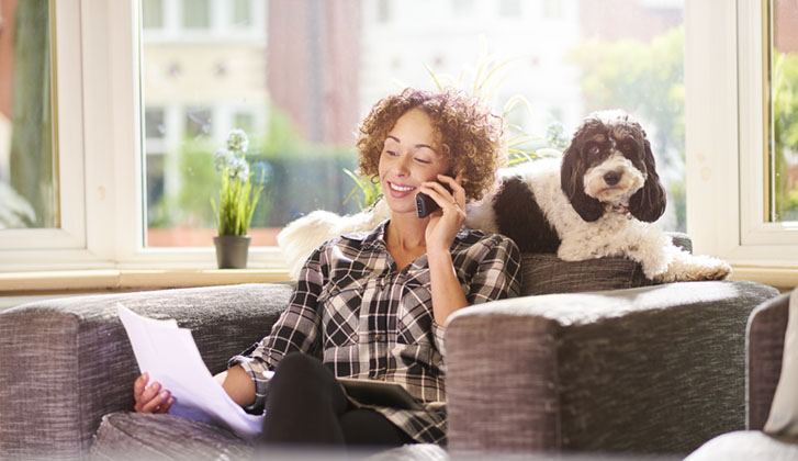 A woman sits on her chair in the living room of her home and checks a bill that she has received . She has her digital tablet on her lap ready to check against her bank account . On the back of her chair her pet dog is relaxing