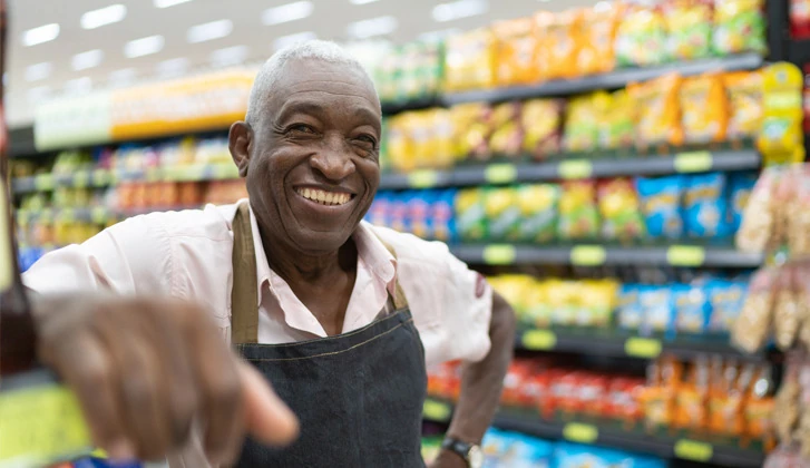 Elderly convenience shop owner, smiling in an aisle.