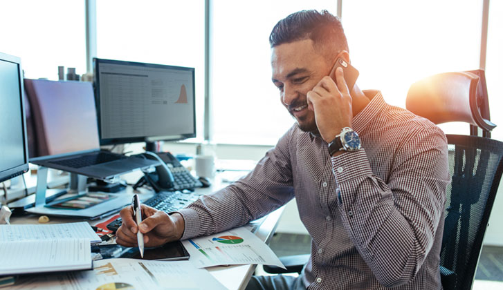 Man in collared dress shirt and watch smiles as he talks on the phone and holds a pen over papers featuring pie charts on a desk with multiple computer screens.