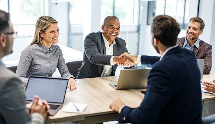 Business men and women seated around an office table while two men shake hands