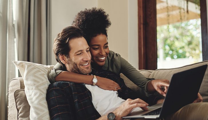 Smiling couple laying on the couch together, looking at a laptop.