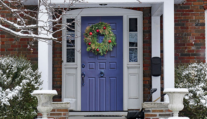 Image of a front door during wintertime. There is a wreath on the door with red ornaments, and snow on the ground.