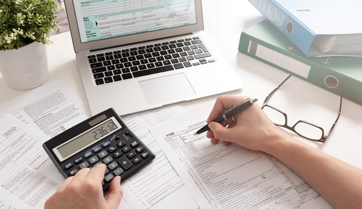 desk view of a person using a calculator a pen and a laptop to file their taxes
