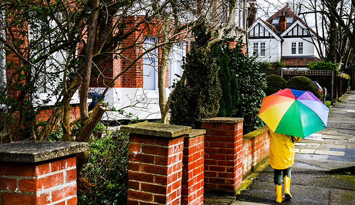 Person with a rainbow umbrella, yellow raincoat, and yellow rainboots walking through a neighborhood.
