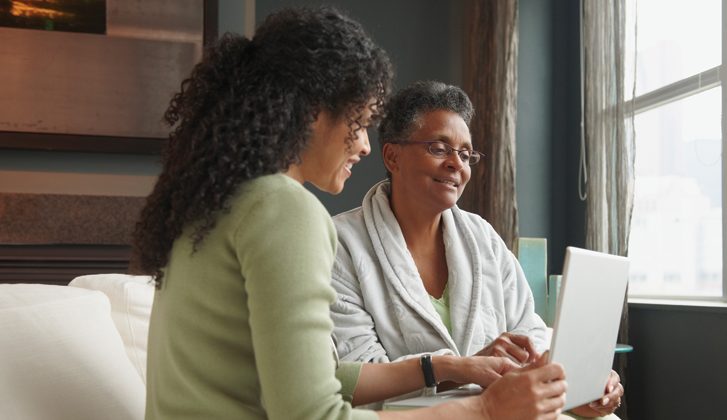 Woman and her elderly mother look at a laptop together, while seated on a couch.