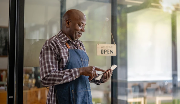 older man standing before an open business sign looking down at tablet screen smiling