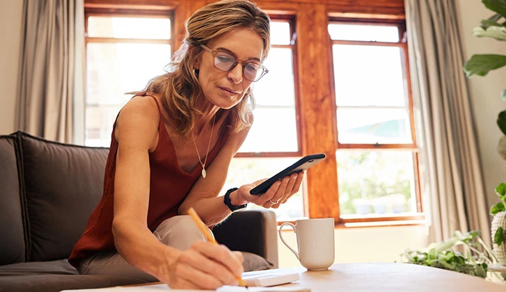 older caucasian woman leaning down toward her coffee table holding a phone in her left hand and writing in a journal with her right hand.
