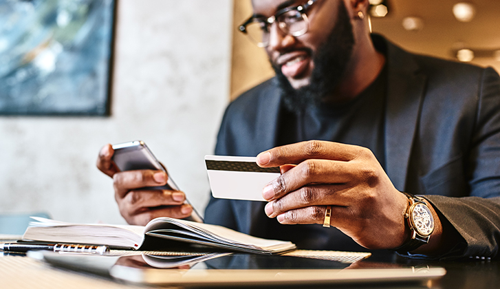 Portrait of African-American male holding cell phone in one hand and credit card in other, making transaction, using mobile banking app during lunch at cafe. His working papers and tablet pc are on the table. Online payment and shopping concept.