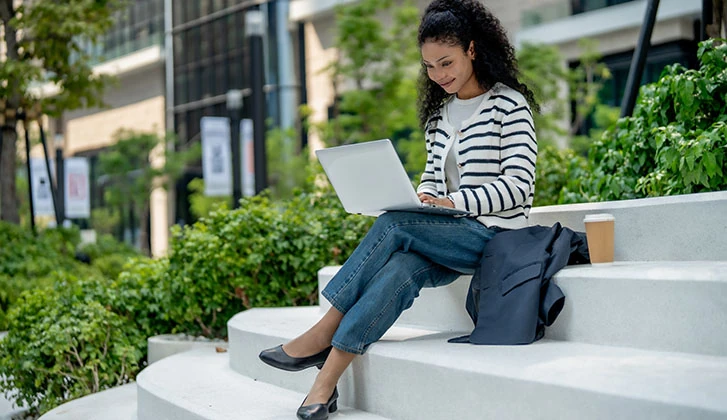 A woman with curly hair sits on outdoor steps, working on laptop. She wears striped sweater and jeans, with coffee cup beside her, surrounded by greenery