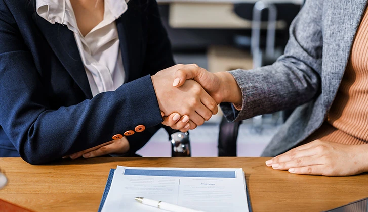 Two business professionals shaking hands over a desk with a contract and pen in front of them.