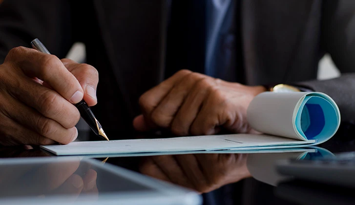 Close-up of a person signing a check with a pen, with a checkbook resting on the desk.