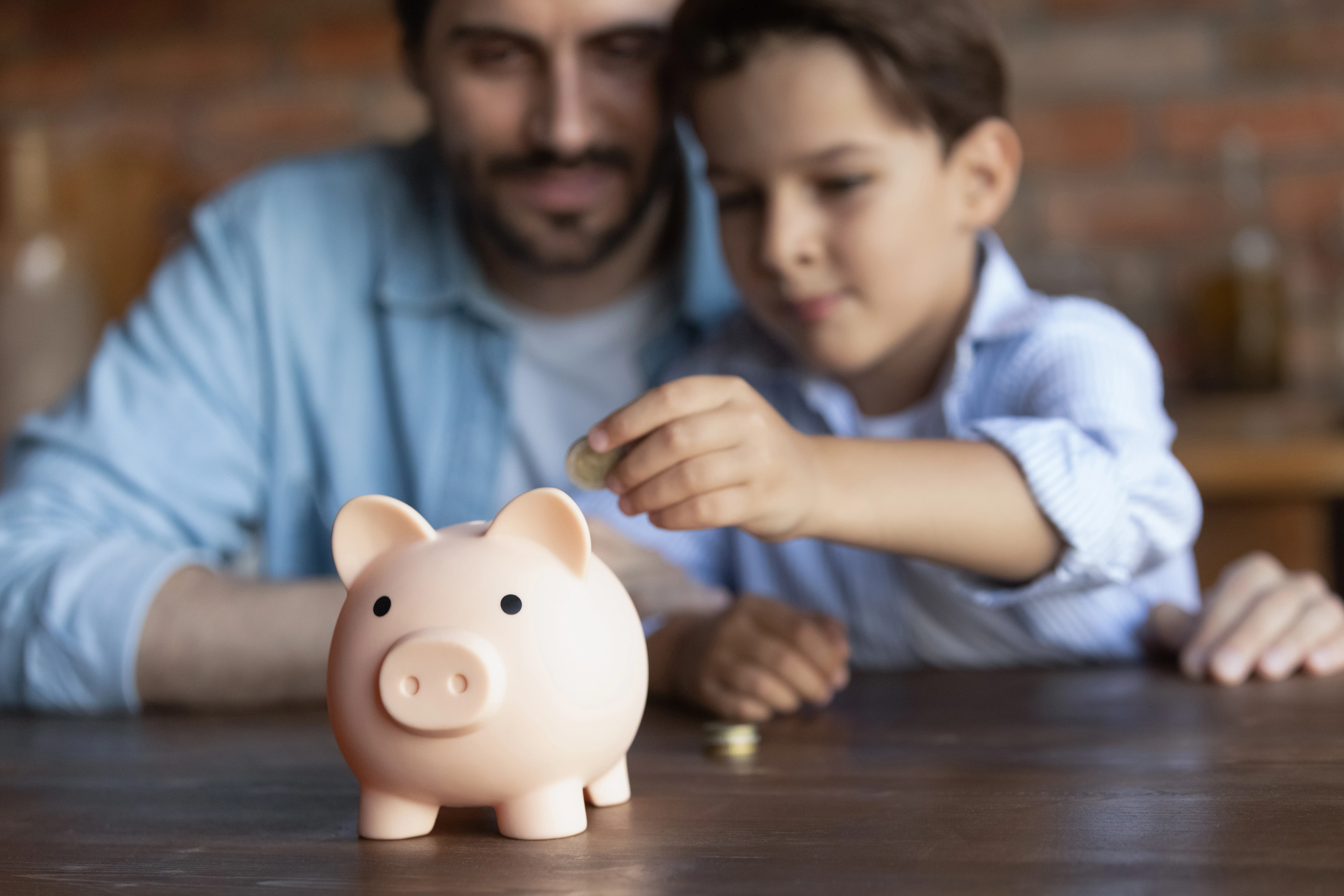 A child is placing a coin into a pink piggy bank while an adult watches closely.