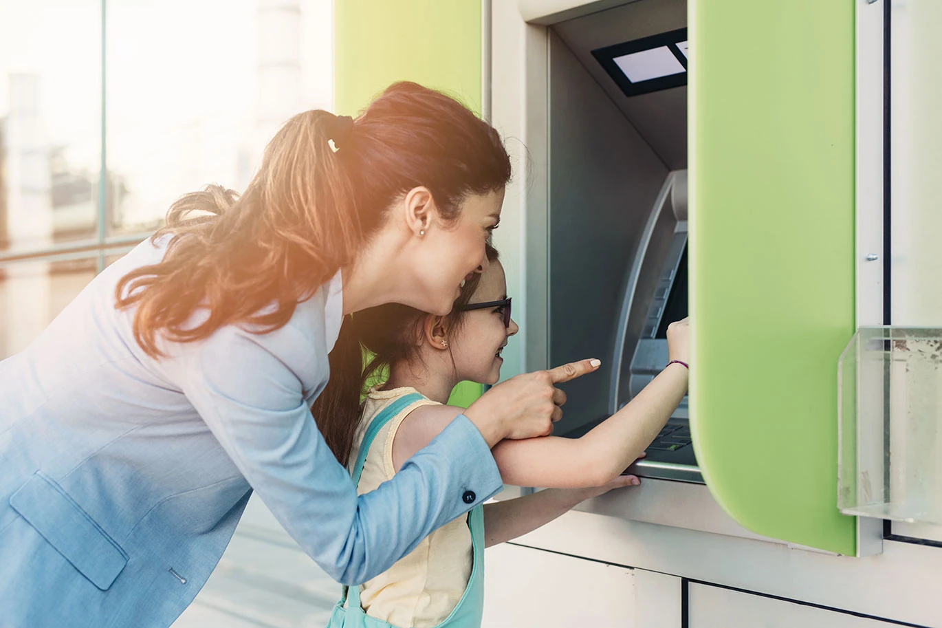 A mother and her young daughter smile as they use an ATM together, with the mother pointing at the screen while the daughter’s hand is on the machine.