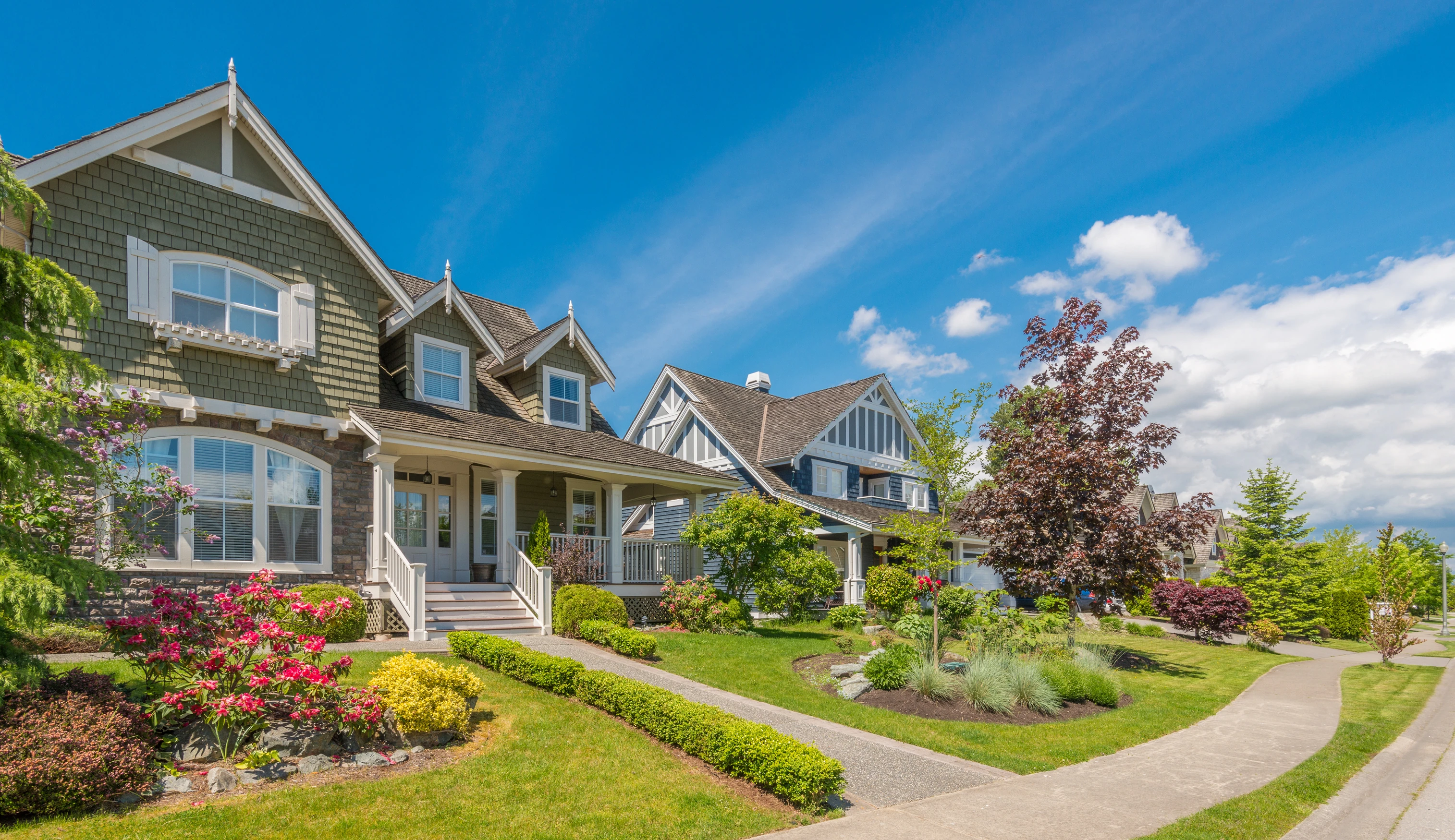 A large family home with green shingles and stone siding sits on a slightly elevated, well manicured lawn in a residential neighborhood.